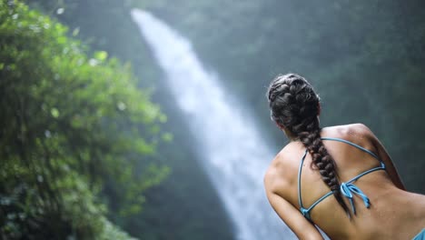 Slow-motion-panning-shot-of-a-girl-in-a-blue-bikini-sitting-in-front-of-a-gushing-NungNung-Waterfall-in-Bali,-Indonesia