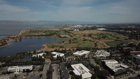 Aerial-birds-eye-view-of-shoreline-lake-Google-campus-amphitheater-park-mountain-background-pan-left