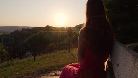 a-woman-in-a-red-dress-looks-at-sunset-sitting-on-a-bench