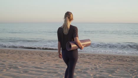 Travel-girl-walking-towards-calming-ocean-on-beach-with-a-yoga-mat