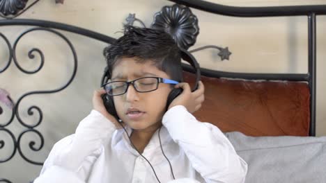 Kleiner-Indischer,-Asiatischer,-Kaukasischer-Junge,-Der-Eine-Brille-Trägt-Und-Zuhört-Und-Musik-über-Kopfhörer-Genießt,-Die-Augen-Schließt-Und-Die-Hände-Am-Ohr-Sitzt-Und-In-Der-Seitenansicht-Des-Wohnzimmers-Sitzt