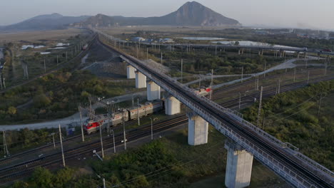 Locomotoras-De-Trenes-Rusos-Que-Pasan-Bajo-El-Puente-Del-Camión-Con-Montañas-En-El-Fondo-De-La-Puesta-Del-Sol,-Federación-Rusa