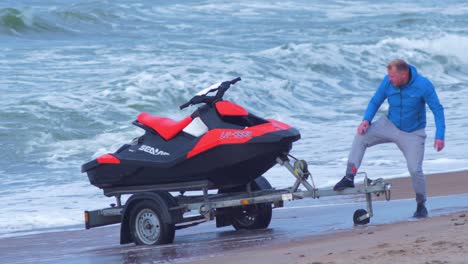 Father-and-daughter-standing-near-a-red-jet-ski-on-the-trailer-on-the-Baltic-sea-sandy-beach,-medium-shot