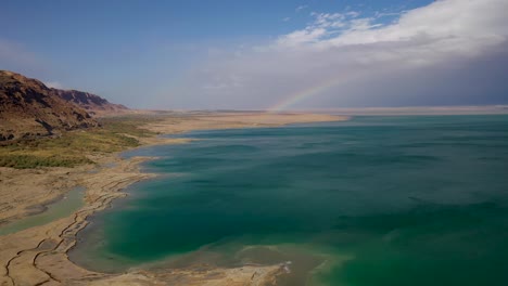 Rainbow-over-green-water-Dead-Sea,-half-cloudy-sky,-forward-fly-drone-shot
