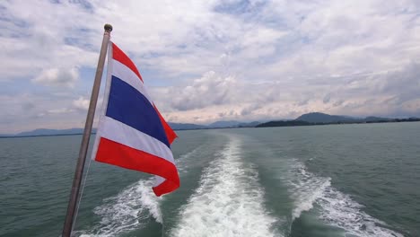 Die-Thailändische-Flagge-Weht-Im-Wind-Auf-Einer-Bootsfahrt-Zur-Insel-Phi-Phi-Mit-Phuket-Im-Hintergrund,-Während-Das-Kielwasser-Des-Bootes-In-Zeitlupe-Durch-Das-Meerwasser-Schiebt