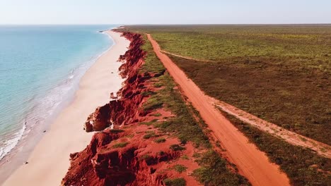 Drone-tracking-dirt-road-atop-red-ocean-side-cliffs-with-sandy-beach-and-blue-water