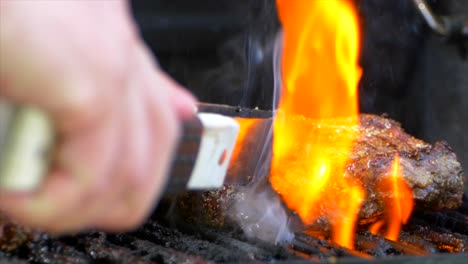 A-pair-of-meat-tongs-slide-a-nearly-cooked-juicy-rib-eye-steak-on-a-grill-and-presses-on-it-as-flames-shoot-up-in-slow-motion