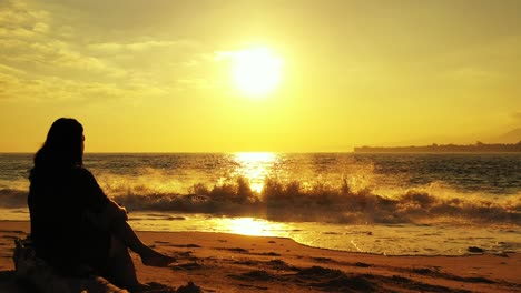 Silhouette-of-woman-sitting-on-quiet-beach-at-sunset-with-glowing-yellow-sky-over-troubled-sea-with-waves-splashing-on-sand,-Vietnam