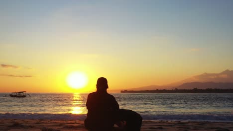 Young-woman-meditating-sitting-on-quiet-exotic-beach-at-magical-sunset-with-yellow-sun-on-bright-sky,-silhouette-of-tropical-island,-Philippines