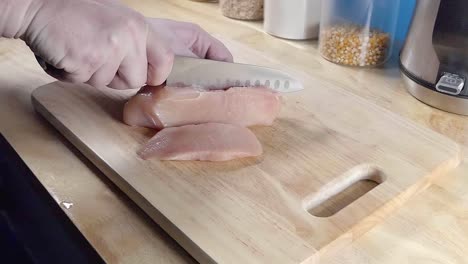 Slow-Motion-Slider-Shot-of-Slicing-Chicken-Breast-Pieces-with-a-Chef's-Knife-on-a-Wooden-Cutting-Board-in-a-Home-Kitchen