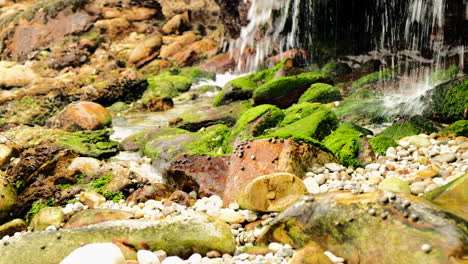 Water-from-stream-falls-on-green-mossy-rocks-on-coastline,-panning-shot