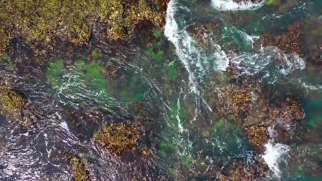 Atemberaubende-Luftaufnahme-Von-Oben-Auf-Wellen,-Die-Auf-Felsen-In-Einem-Tropisch-Grün-Aussehenden-Meer-Schlagen