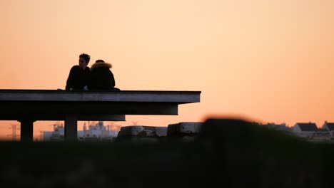 Young-couple-sitting-on-the-deck-enjoying-the-warm-sunset-with-city-in-background