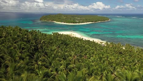 Aerial-shot-flying-over-the-top-of-the-palm-trees-in-the-jungle,-revealing-a-beautiful-and-quiet-white-sand-beach-with-a-small-island-nearby-in-the-secluded-paradise-with-a-few-small-boats