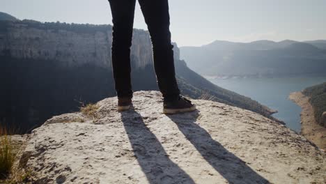 Scenic-view-of-mountain-ranges-and-lake-in-background-with-close-up-low-angle-view-of-man's-legs-standing-dangerously-at-edge-of-cliff-observing-landscape,-handheld-closeup
