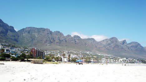Panning-view-of-Camps-Bay-from-the-white-sandy-beaches-looking-at-the-apartments,-and-resorts-with-the-Twelve-Apostles-mountains-in-the-background