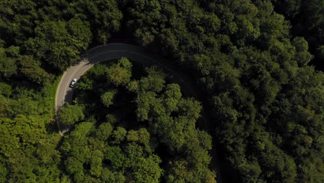 Aerial-view-at-a-white-car-from-above-drining-into-a-tight-curve-in-a-forest,-look-up-shot-to-a-curvy-river-under-the-blue-skyed-horizin-at-summertime