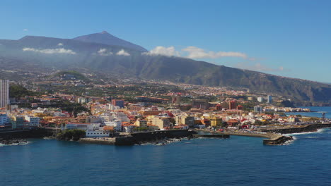Scenic-view-of-the-city-of-Puerto-de-la-Cruz,-Canary-Islands,-Spain,-its-harbor-on-the-coast-of-the-Atlantic-ocean,-below-the-Pico-de-Teide-mountain,-blue-sea-and-sky,-zooming-aerial-shot-4K
