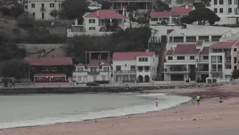 People-At-The-Beach-In-Sao-Martinho-do-Porto-In-Leiria,-Portugal---Beautiful-Village-Houses-In-The-Background---static-shot,-slow-motion
