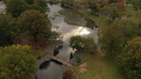 An-aerial-drone-shot-over-a-park-in-the-day-with-a-pond,-with-an-arched-footbridge