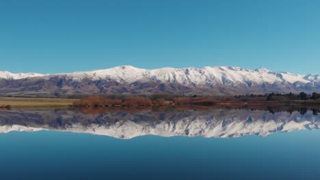 Flock-of-white-birds-flying-over-a-still-lake,-with-a-perfect-reflection-of-the-mountain-range-and-blue-sky