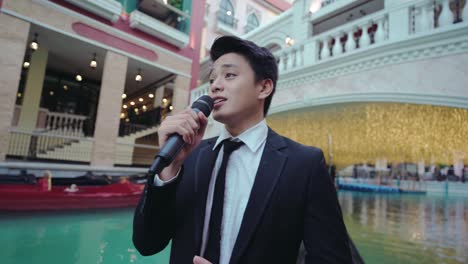 Young-Man-In-Black-Suit-Holding-A-Microphone-Singing-While-Riding-The-Gondola-At-Venice-Grand-Canal-In-Mckinley-Hill,-Taguig-City,-Metro-Manila,-Philippines