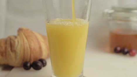 Pouring-fresh-orange-juice-at-breakfast-with-honey-and-croissants