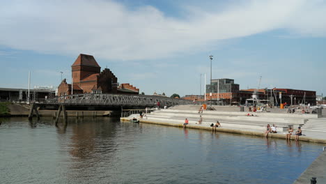 People-sitting-on-waterfront-beside-old-bridge-in-Lübeck-during-summertime,wide-shot