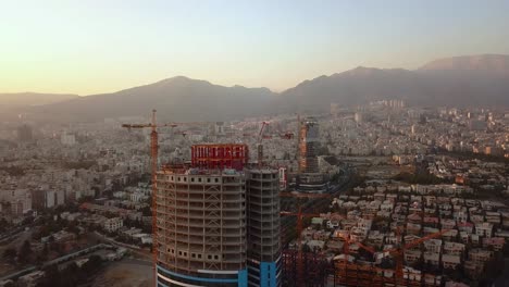 Rotating-Around-Tower-in-Construction-Building-Drone-Aerial-Shot-in-Tehran-Iran-Cityscape-Buildings-in-Background-in-Sunset-Time-and-Mountain-in-Landscape-and-Air-Pollution-Visible-in-Horizon
