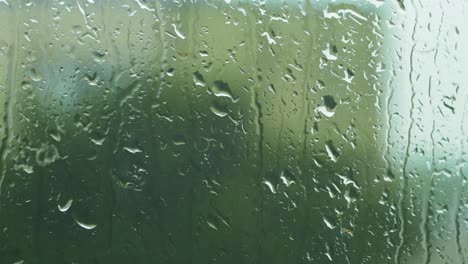 Rain-drops-sliding-slow-on-window-glass-in-rainy-day,-de-focused-tree-in-background,-medium-closeup-shot
