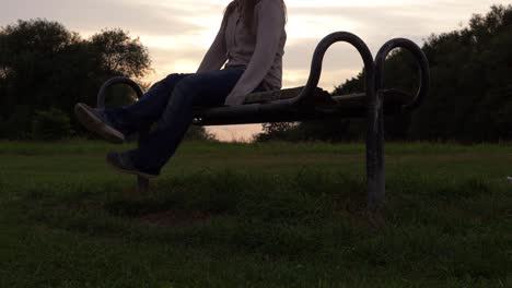 Legs-and-feet-of-woman-sitting-on-a-park-bench-at-sunset