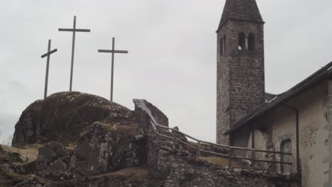 camera-tilts-up-towards-a-small-rock-church-with-three-crosses-alongside,-in-a-wood-of-the-Italian-Alps-4k-slow-motion-bell-tower-detail