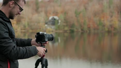 Man-walking-over-to-the-tripod-with-a-tight-crop-putting-the-camera-on-a-tripod-in-landscape-orientation-with-fall-colours-and-a-lake-in-the-background