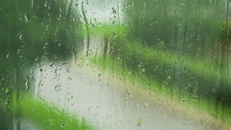 An-Einem-Regnerischen-Tag-Gleiten-Regentropfen-Langsam-über-Die-Fensterscheibe,-Unscharfe-Straße-Mit-Fahrendem-Auto-Im-Hintergrund,-Mittlere-Nahaufnahme