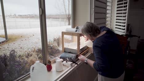 Mature-woman-adds-water-to-her-indoor-hydroponics-garden---snowy-day-outside-her-basement-window