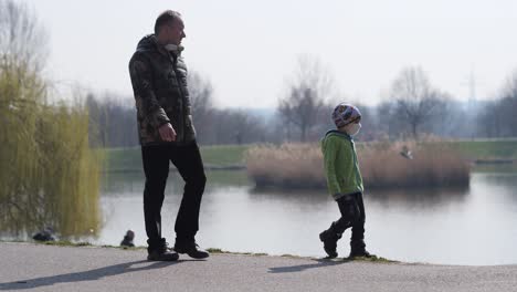Child-With-Face-Mask-Walking-in-Park-by-Lake-With-Father-on-Sunny-Morning