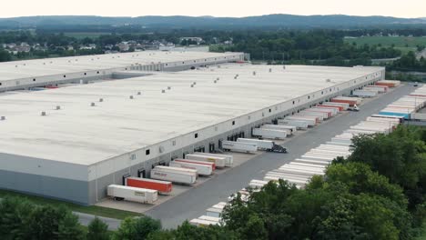 Aerial-revealing-shot-of-large-distribution-center,-warehouse,-shipping-depot,-trucks-load-and-unload-packages-at-dock,-logistics-theme
