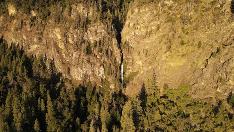 Aerial-rising-over-Corbata-Blanca-thaw-waterfall-hiding-between-mountains-surrounded-by-pine-trees-at-sunset,-Patagonia-Argentina