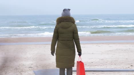 Young-woman-walking-along-the-beach-in-winter-overcast-day,-red-bag-in-right-hand,-handheld-medium-shot