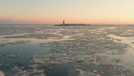 Breakwater-and-lighthouse-at-sunset-with-icy-sea,-tracking-drone-shot