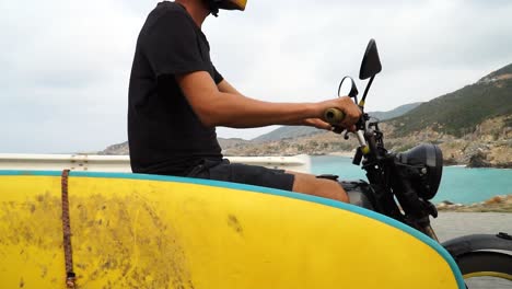 Young-man-drives-motorcycle-with-yellow-surfboard-attached-looking-for-ideal-spot-for-water-kitesurfing