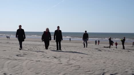 A-group-of-fully-clothed-people-walking-in-a-beach-in-Belgium,-slow-wide-shot
