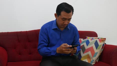 Ein-Asiatischer-Geschäftsmann-Erhält-Gute-Nachrichten-Von-Seinem-Handy,-Während-Er-Auf-Dem-Roten-Sofa-Sitzt