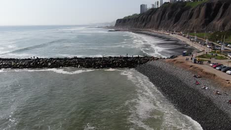 4k-daytime-aerial-video-looking-over-the-Pacific-ocean-hitting-with-power-the-cobblestone-beaches-of-Lima,-Peru,-Miraflores-west-coast,-looking-over-a-peer-of-stones-with-people