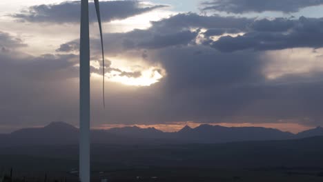 Mountain-range-in-the-distance-with-sun-setting-behind-storm-clouds-with-slow-turning-wind-turbine-blade-in-the-foreground
