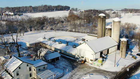 AERIAL-Snow-Falling-Over-Farm-Property-Buildings,-Fields-Of-White-Ground-Cover