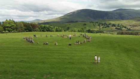 Ancient-site-of-Castlerigg-Stone-Circle-aerial-rising-view-showing-mountains-in-background-and-people-enjoying-themselves,-during-summertime-Keswick-Cumbria-UK