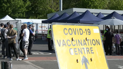 Coronavirus,-COVID-19-Vaccination-Centre-Sign-with-members-of-the-public-and-officials-in-the-background