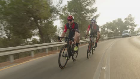 Dos-Ciclistas-De-Carretera-Profesionales-Cabalgando-Por-La-Carretera-Vacía-Del-Bosque