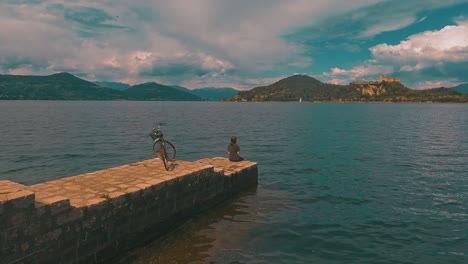 Back-view-of-lonely-woman-with-bicycle-behind-sitting-on-pier-edge-on-Maggiore-lake,-Italy